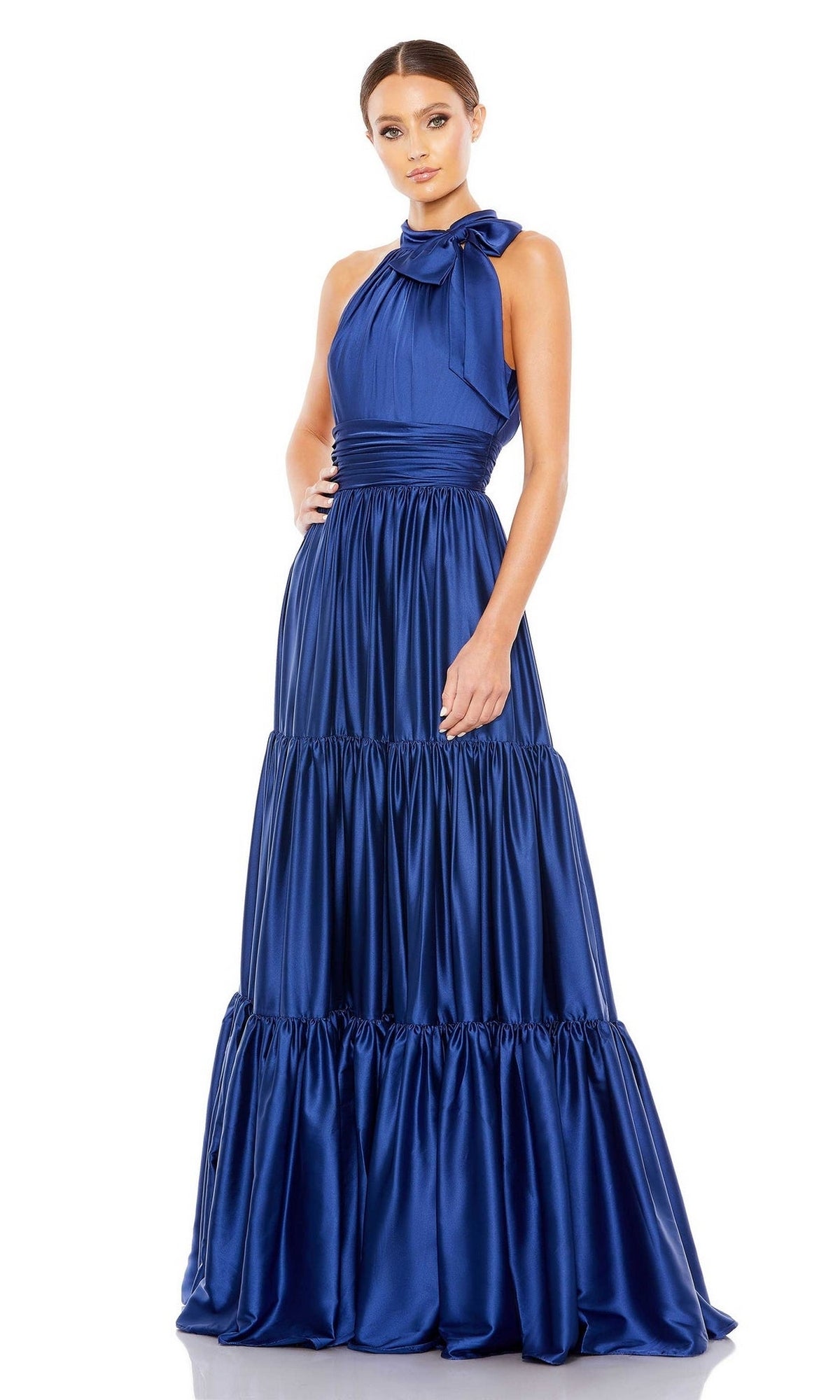 A woman wears the Mac Duggal High-Neck Satin Formal Dress 506581 in royal blue, a sleeveless, floor-length A-line satin gown with a tiered ruffled skirt and an elegant shoulder bow.